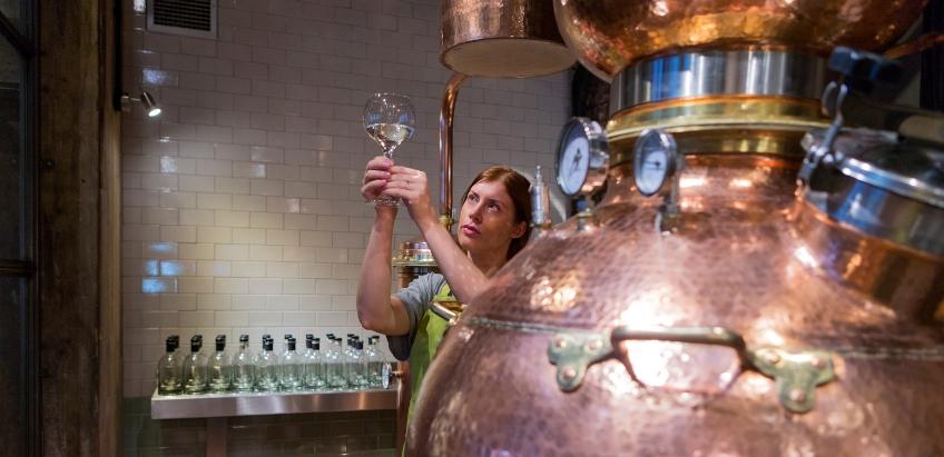 A woman inspecting a glass of gin under the light next to a distillation machine.